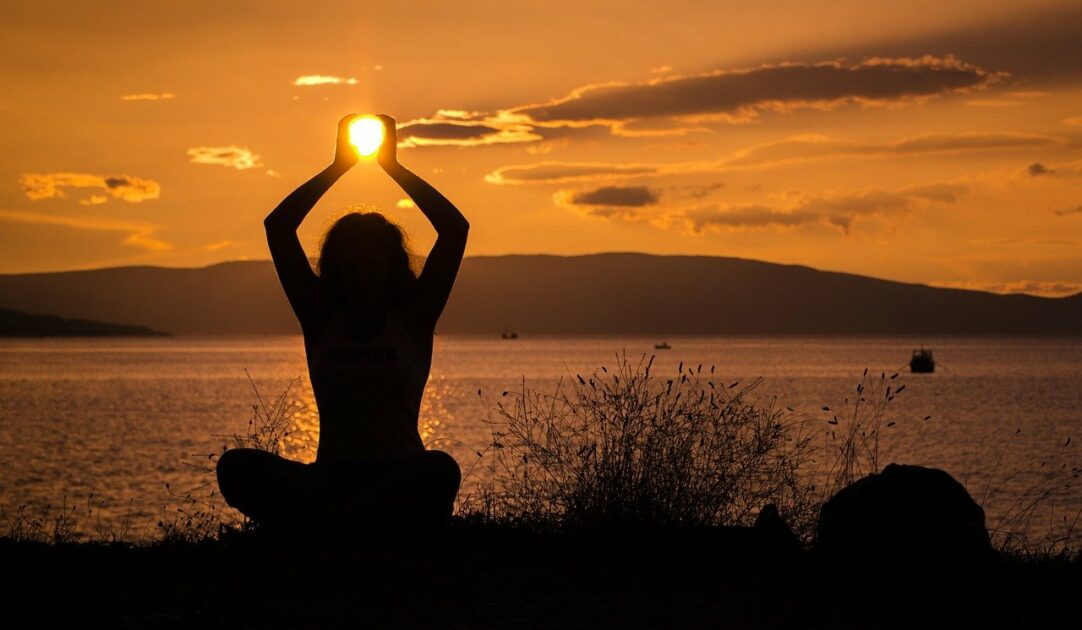Frau übt Yoga am See-Ufer bei Sonnenuntergang. Es wirkt, als hält sie die Sonne in den Händen.